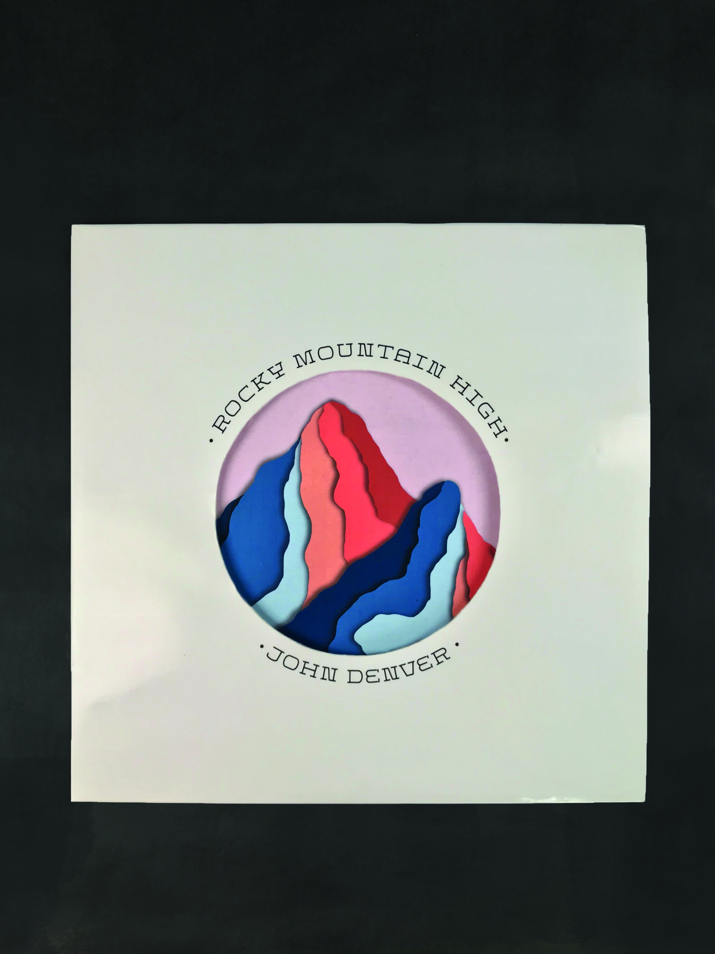 The front cover of my redesign of John Denver's album, Rocky Mountain High. It is a shiny, cream paper with a die-cut of bright, colorful mountains on the sleeve behind it.