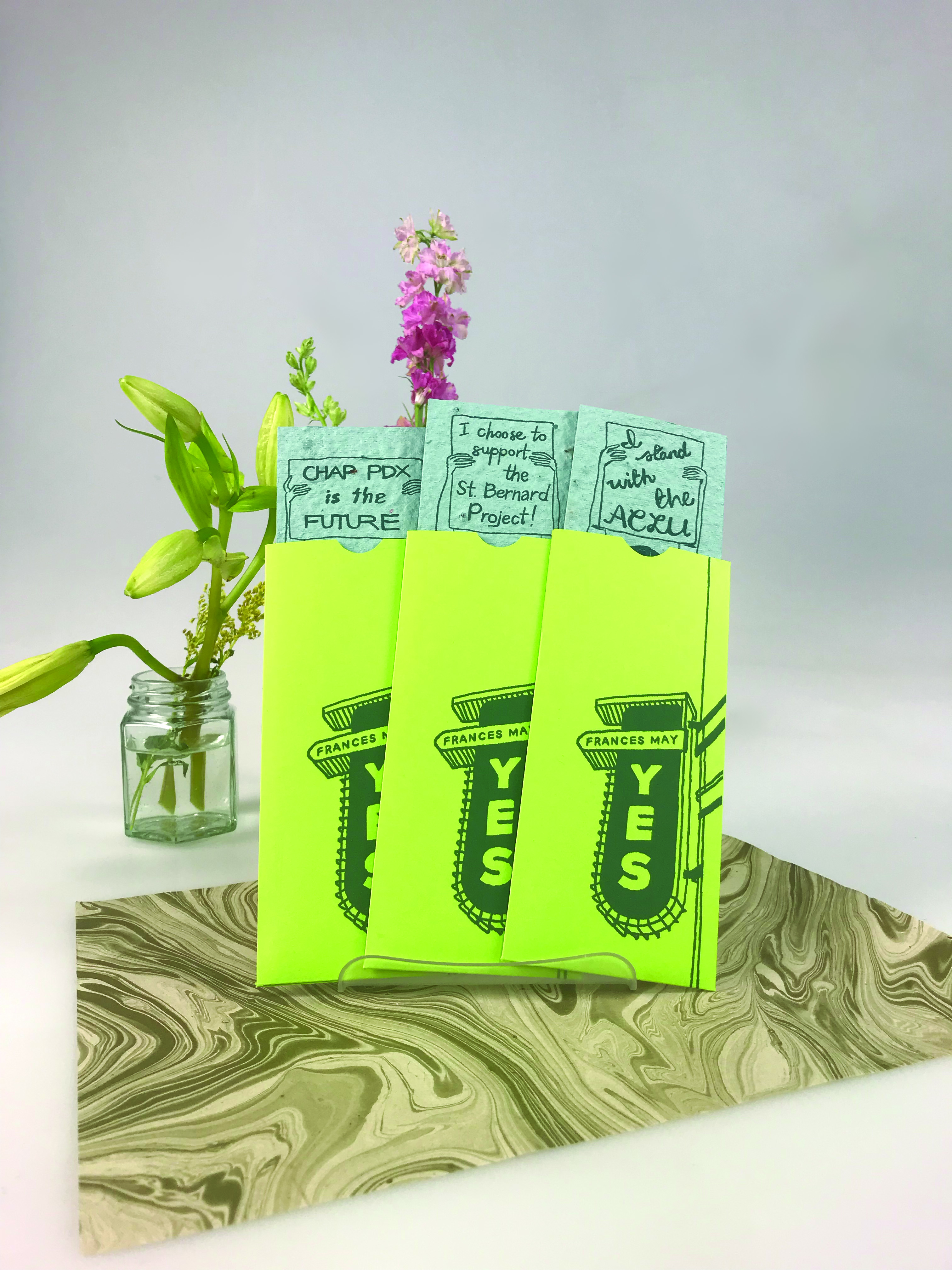 front of bright green gift card sleeves with blue wildflower paper gift cards. The sleeves have an illustration of the Frances May sign, while the cards have illustrations of different women on the front.